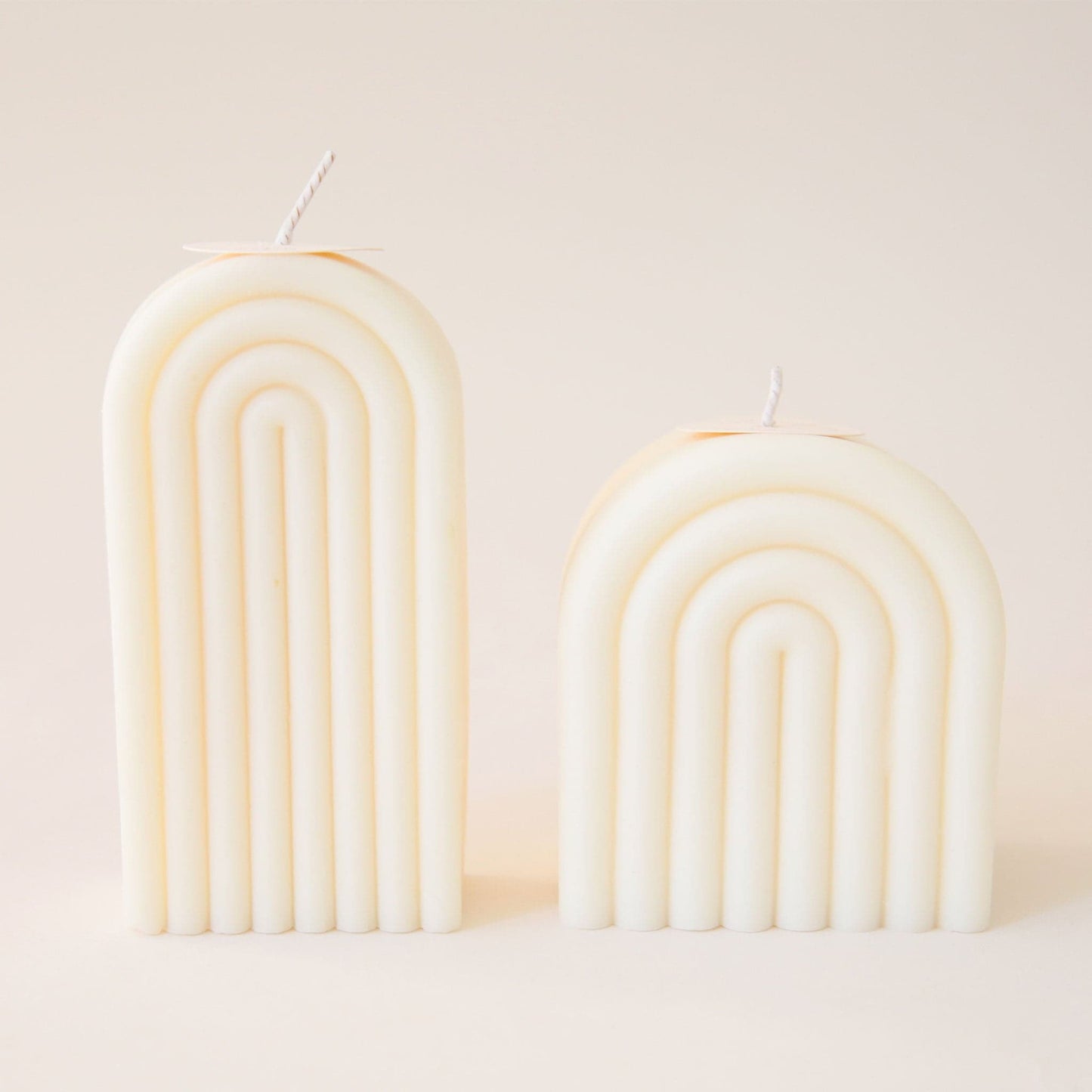Two light cream arch shaped candles, one tall and one short. Each is carved with rainbow-shaped curves and has its own white wick sprouting from the top.