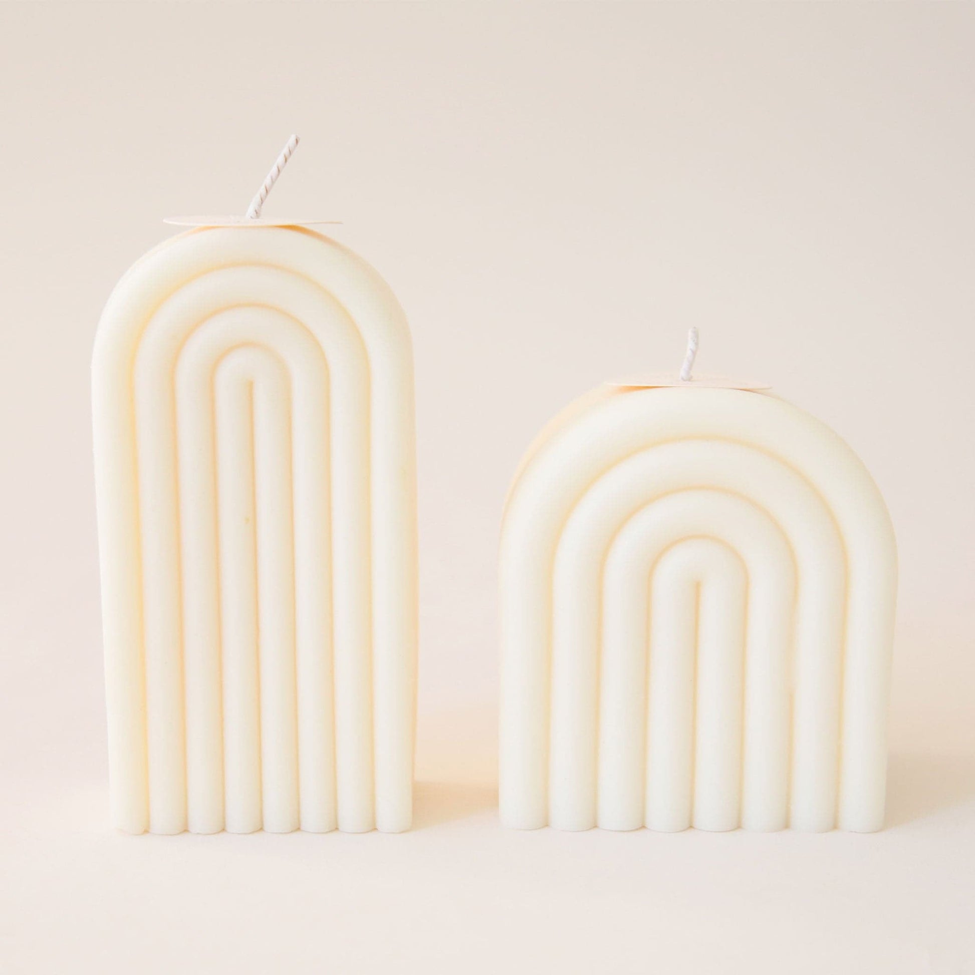 Two light cream arch shaped candles, one tall and one short. Each is carved with rainbow-shaped curves and has its own white wick sprouting from the top.
