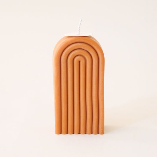 Single tall, arch shaped candle. The candle is carved with rainbow-shaped curves and has a white wick sprouting from the top.